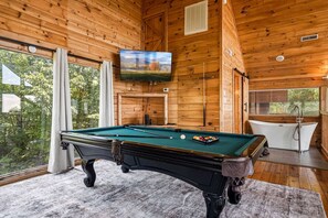 Ein Billardtisch mit grüner Spielunterlage, ein an der Wand befestigtes Fernsehgerät und eine Badewanne in einer Holzhütte mit großen Fenstern.