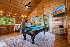 Interior de una cabaña de madera con mesa de billar, ventilador de techo, televisor y ventana con vistas a la vegetación.