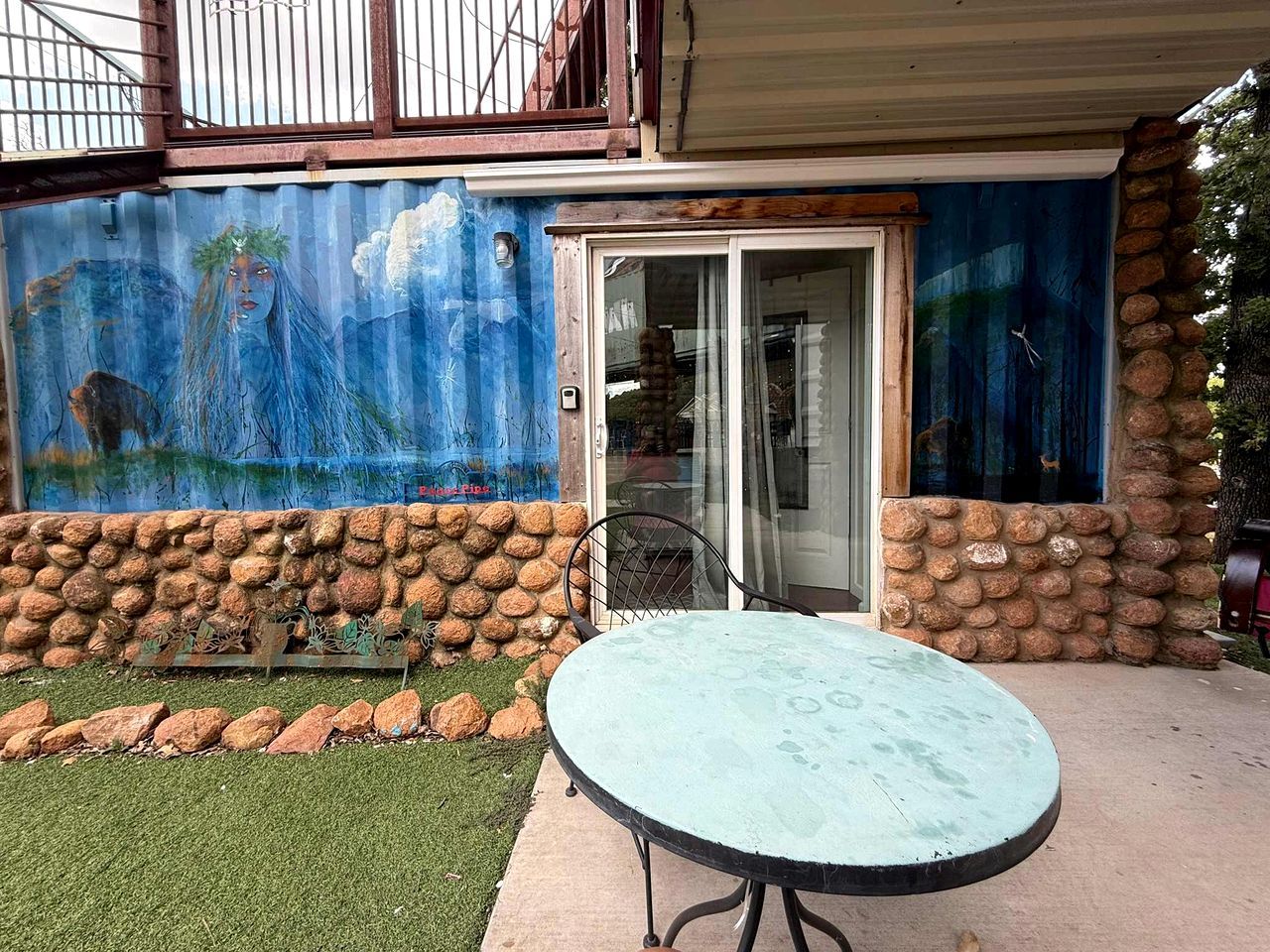 A patio with a round table and chairs, a stone wall, and a mural on the building.