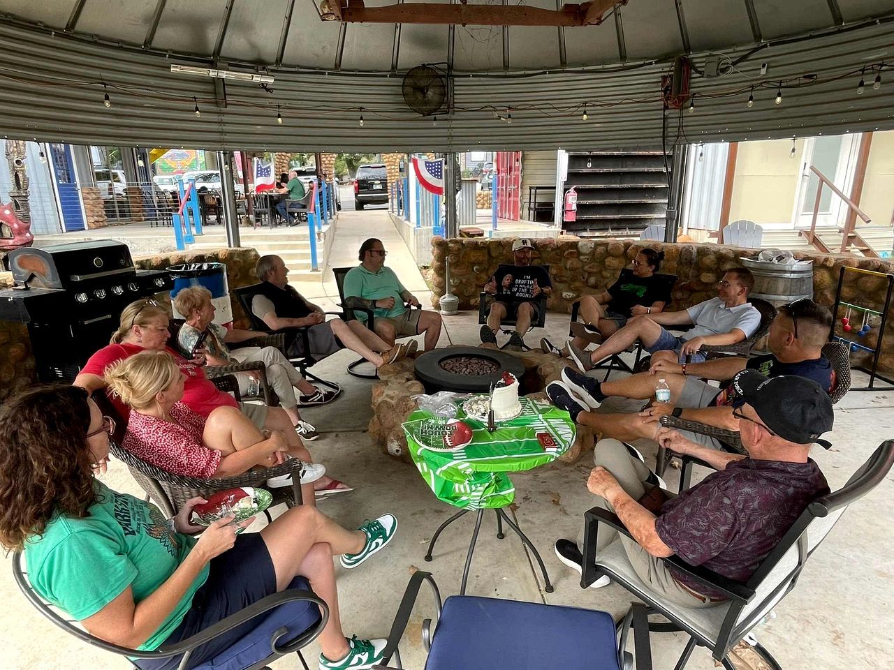 Um grupo de pessoas sentadas à volta de uma mesa com bolo e bebidas, sob uma área coberta com teto de metal.