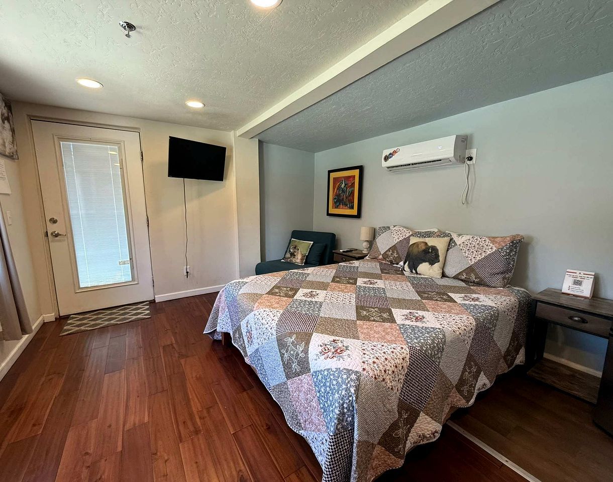 A bedroom with a wooden floor, a bed with a quilt, a TV mounted on the wall, and an air conditioning unit.