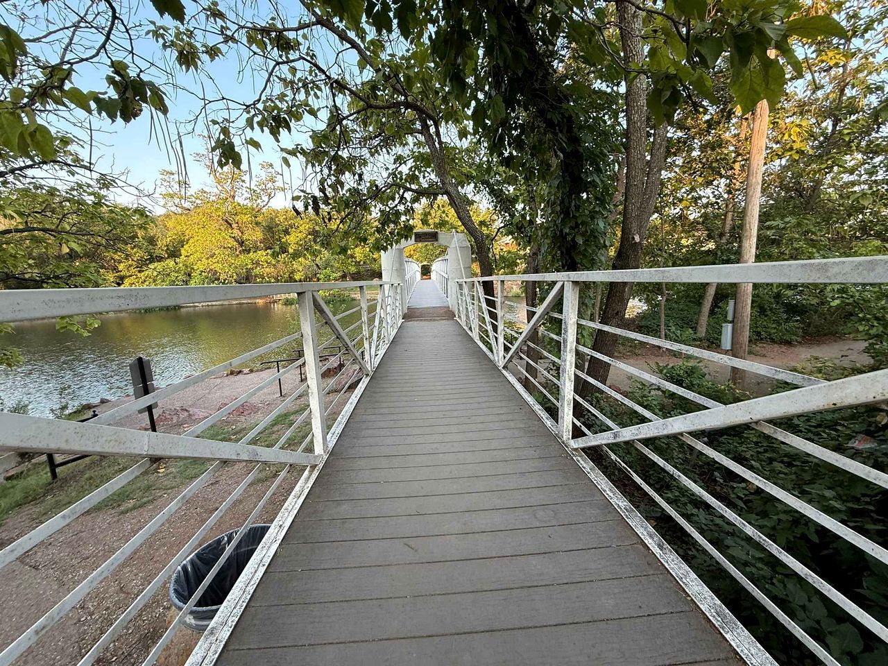 Un puente peatonal de madera con barandillas blancas que cruza un cuerpo de agua, rodeado de árboles.