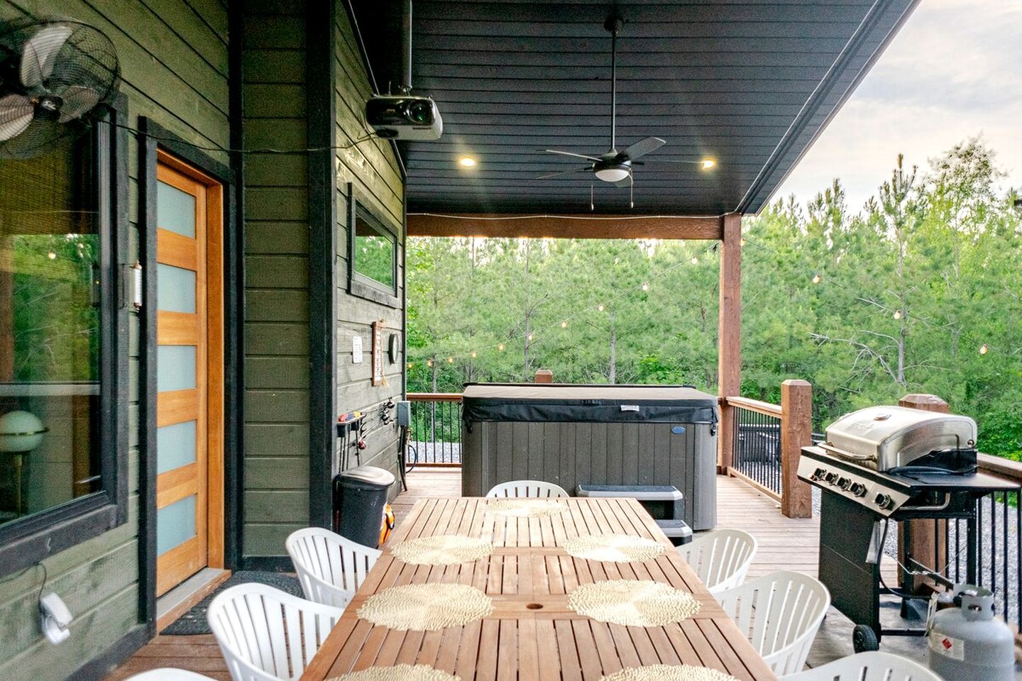 Um deck ao ar livre com mesa de madeira, cadeiras brancas, ventilador de teto e churrasqueira.