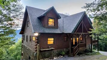 A wooden cabin with a porch, a stone foundation, and a view of the surrounding forest.