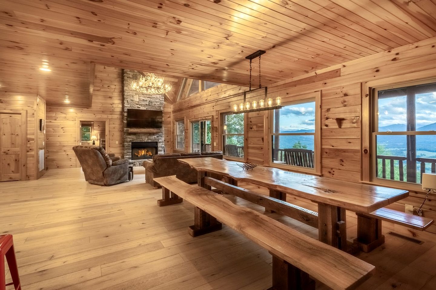 A log cabin interior with a fireplace, wooden benches, and large windows offering a view.