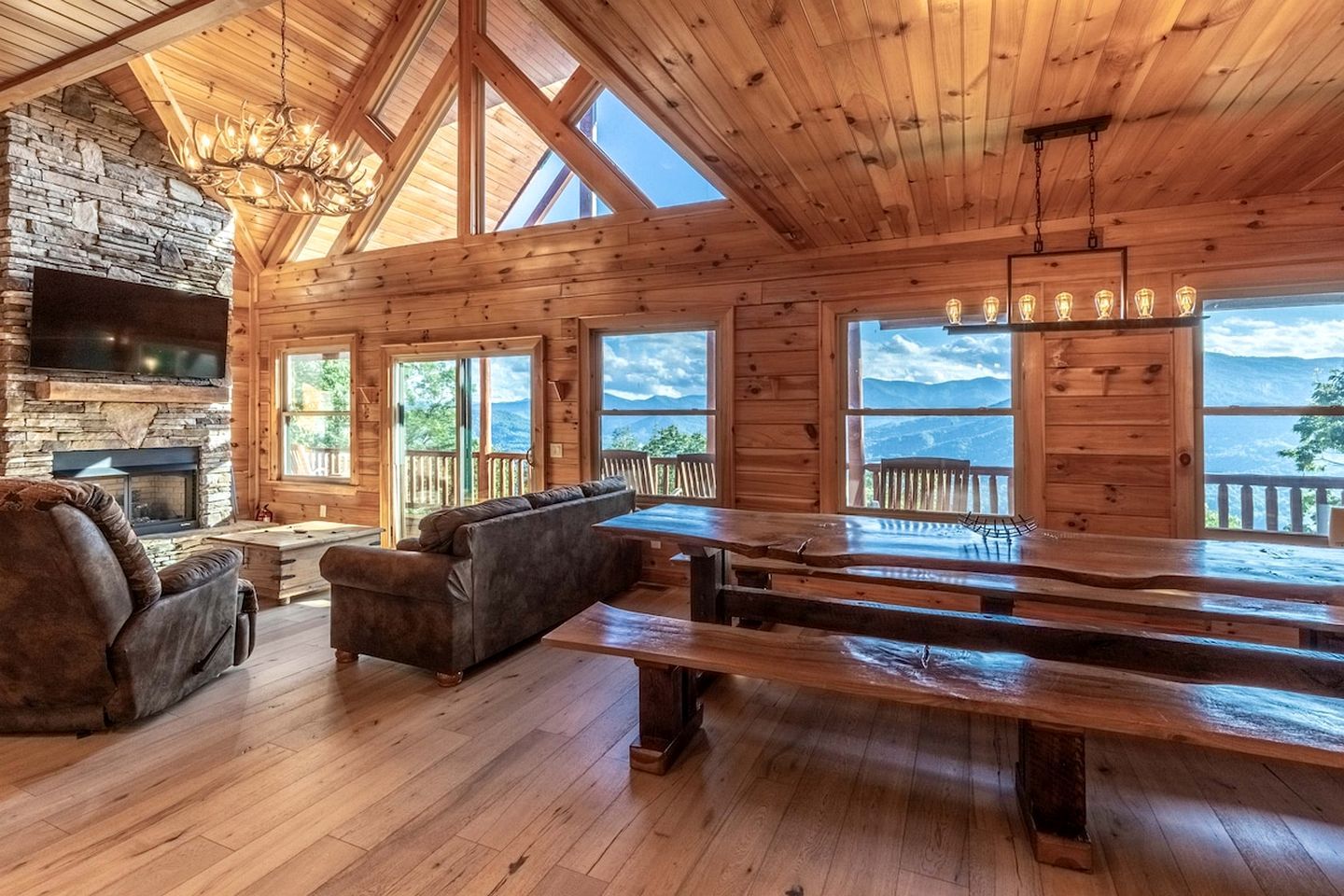 Ein Blockhaushaus-Interieur mit Kamin aus Stein, Holzmöbeln und großen Fenstern mit Blick auf eine gebirgige Landschaft.