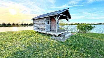 Une cabane en bois dotée d’un porche, située dans une zone herbeuse près d’une étendue d’eau.