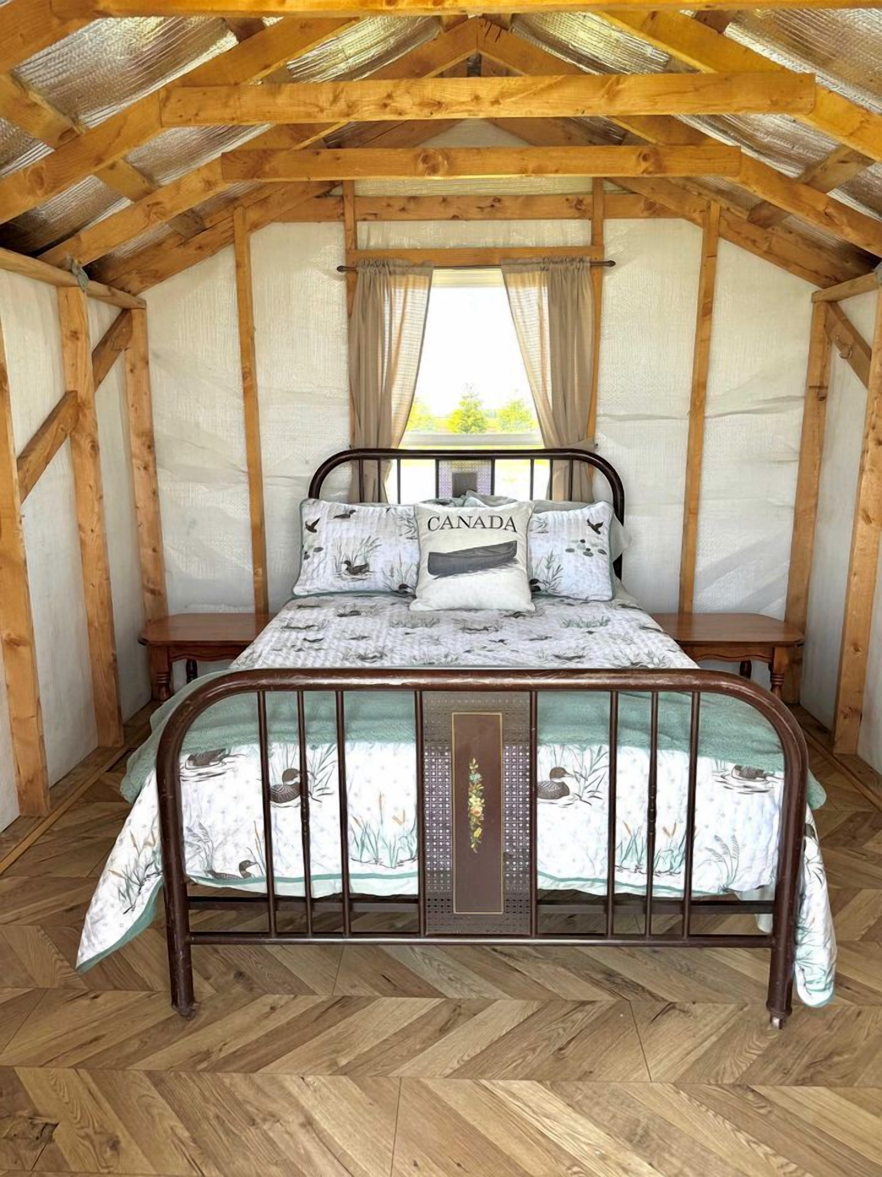 Une pièce aux murs en bois, avec un lit, une fenêtre ornée de rideaux et un parquet à motifs en chevrons.