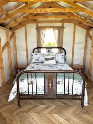 A wooden-framed room with a bed, a window with curtains, and a herringbone-patterned floor.