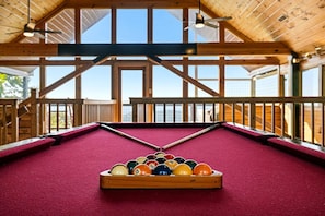 Ein Billardtisch mit Holzrahmen und roter Spielvliesdecke, umgeben von Fenstern und Holzbalken.