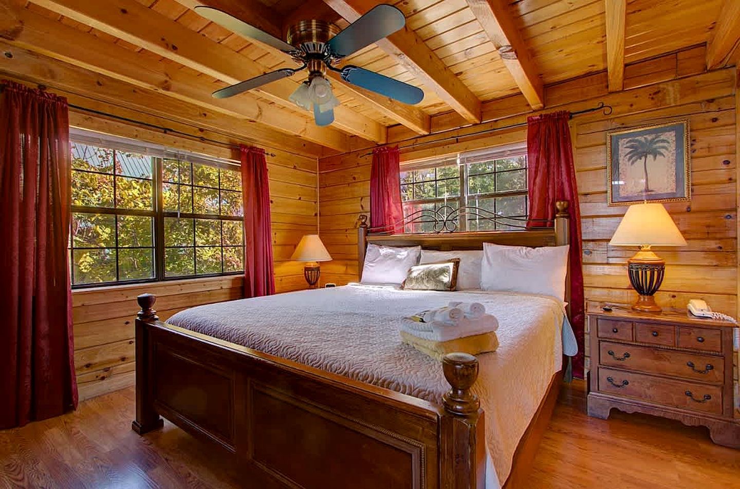 Une chambre à coucher de style cabane en bois, avec un lit, deux lampes de chevet, un ventilateur de plafond et une fenêtre avec des rideaux.