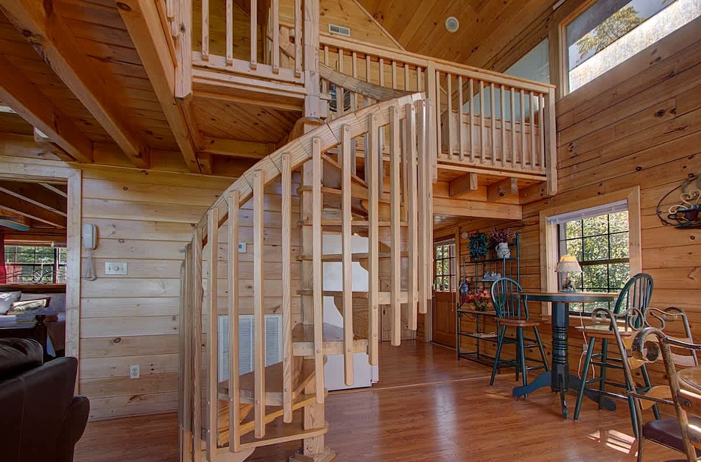 A wooden cabin interior with a spiral staircase, a dining area with chairs and a table, and a kitchen area.