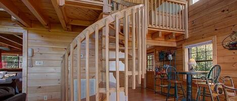 A wooden cabin interior with a spiral staircase, a dining area with chairs and a table, and a kitchen area.
