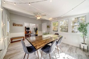 Un coin repas avec une table en bois, quatre chaises, un ventilateur de plafond et de grandes fenêtres.