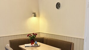 A corner seating area with a wooden table, cushioned bench seating, a vase with red flowers, and a wall-mounted clock.