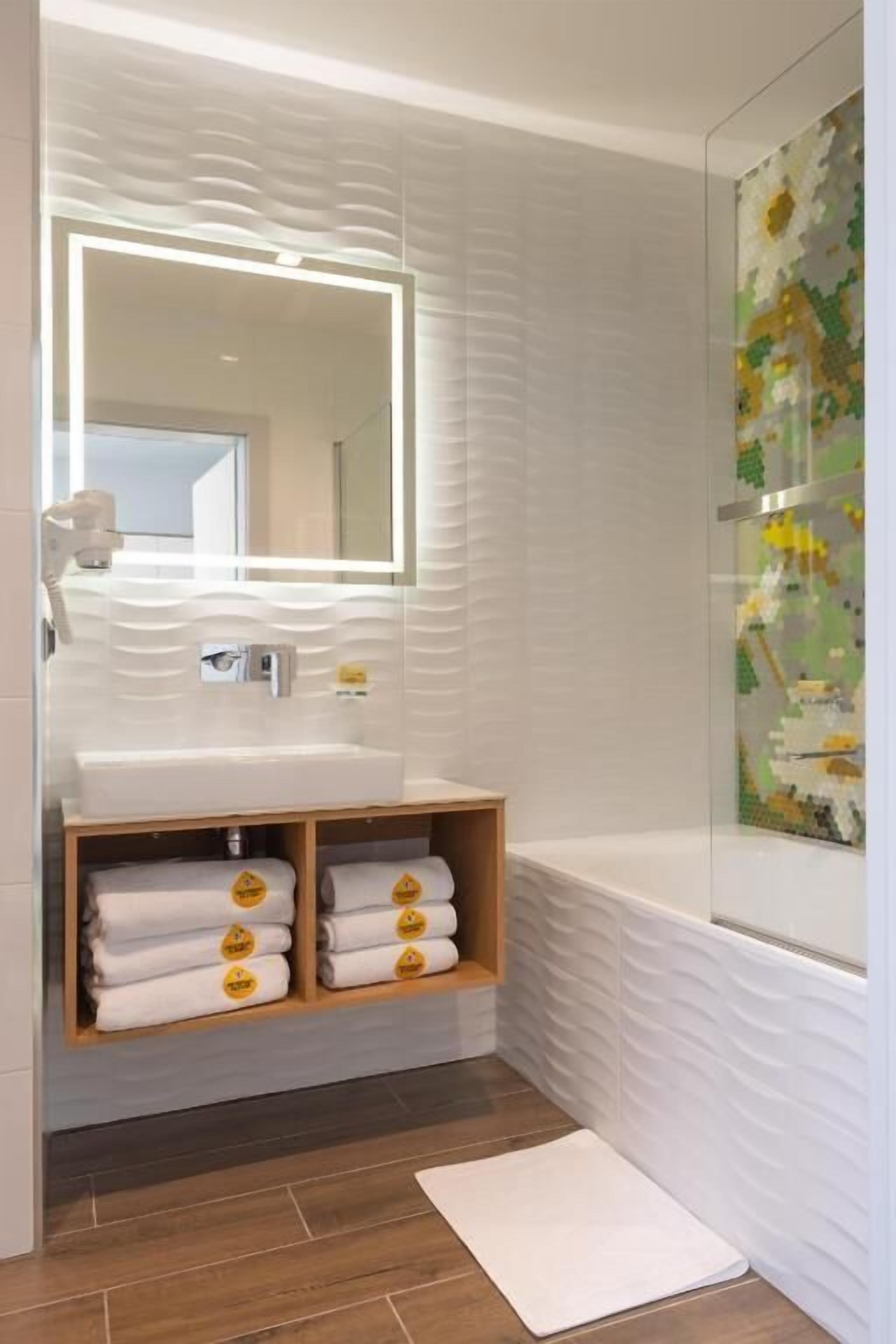 A bathroom with a white sink, a mirror with a light, a wooden shelf with towels, a bathtub, and a tiled floor.