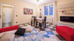 Een kamer met een bed, een rode bank, een eettafel met stoelen, een televisie en een raam met gordijnen.