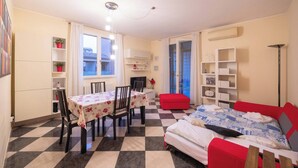 Een kamer met een zwart-wit geruite vloer, een eettafel met stoelen, een bed met rode en witte beddengoed, en een rode poef.