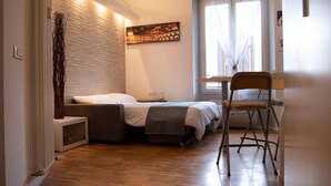 Un dormitorio con cama, una silla, escritorio, ventana con cortinas, radiador y un cuadro en la pared.