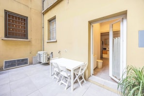 Une terrasse avec une table et des chaises blanches, une porte-fenêtre coulissante donnant sur la cuisine et un petit climatiseur.