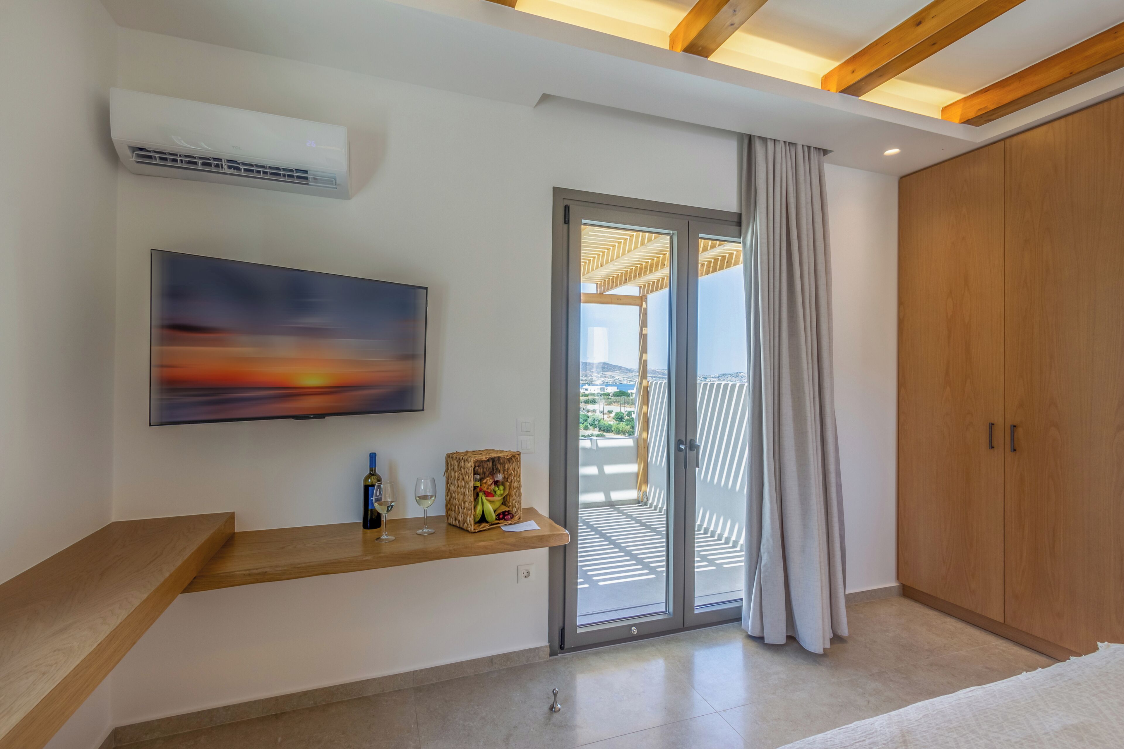 A modern room with a flat-screen TV, a wooden bench, a sliding glass door to a balcony, and a wardrobe.
