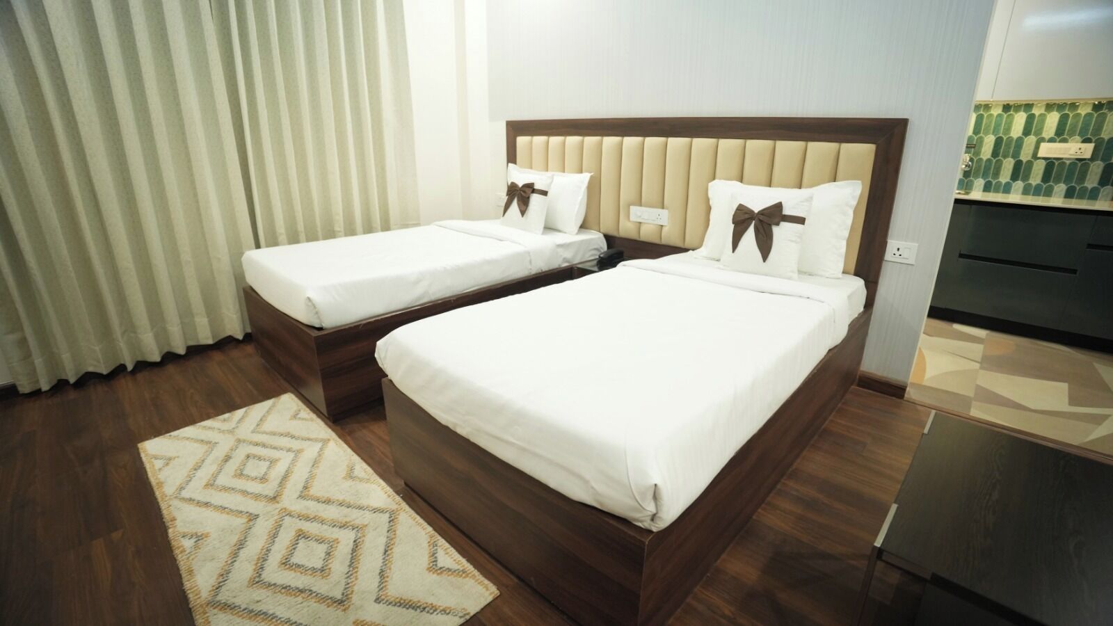 A hotel room with two beds, a wooden floor, a patterned rug, and a kitchen area.