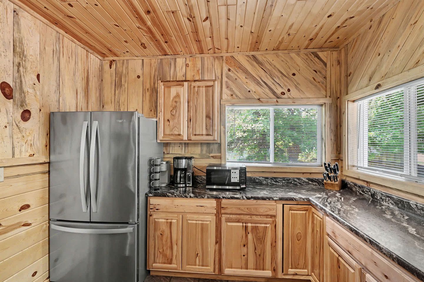 A kitchen with wooden cabinets, a stainless steel refrigerator, a coffee maker, a microwave, and a window with blinds.