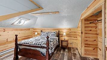 Een slaapkamer met houten lambrisering en een schuine hemel, een bed met een geruit sprei, twee nachtkastjes met lampen, en een dakraam.