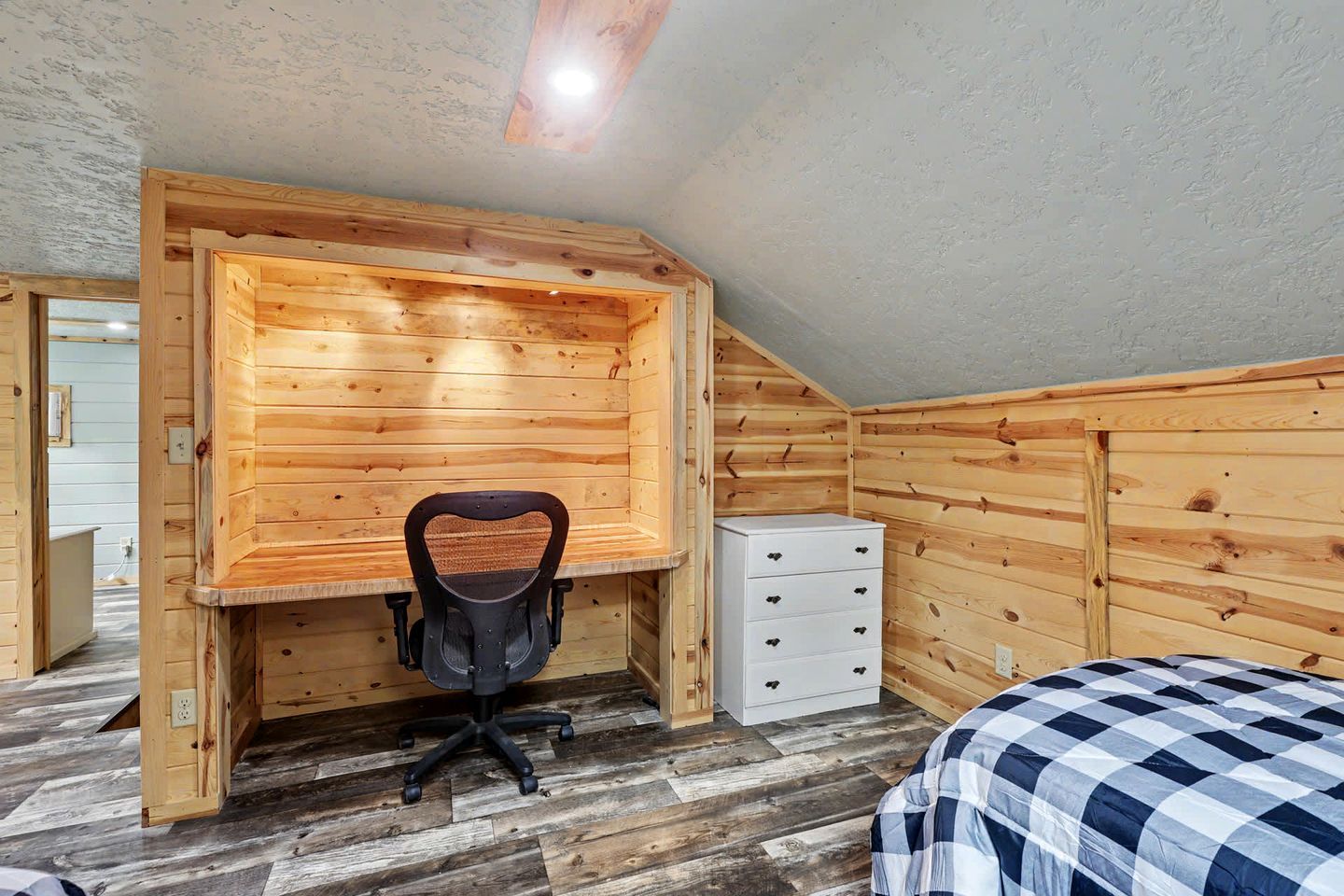 A room with a sloped ceiling, wooden walls, a desk with a chair, a dresser, and a bed with a checkered bedspread.