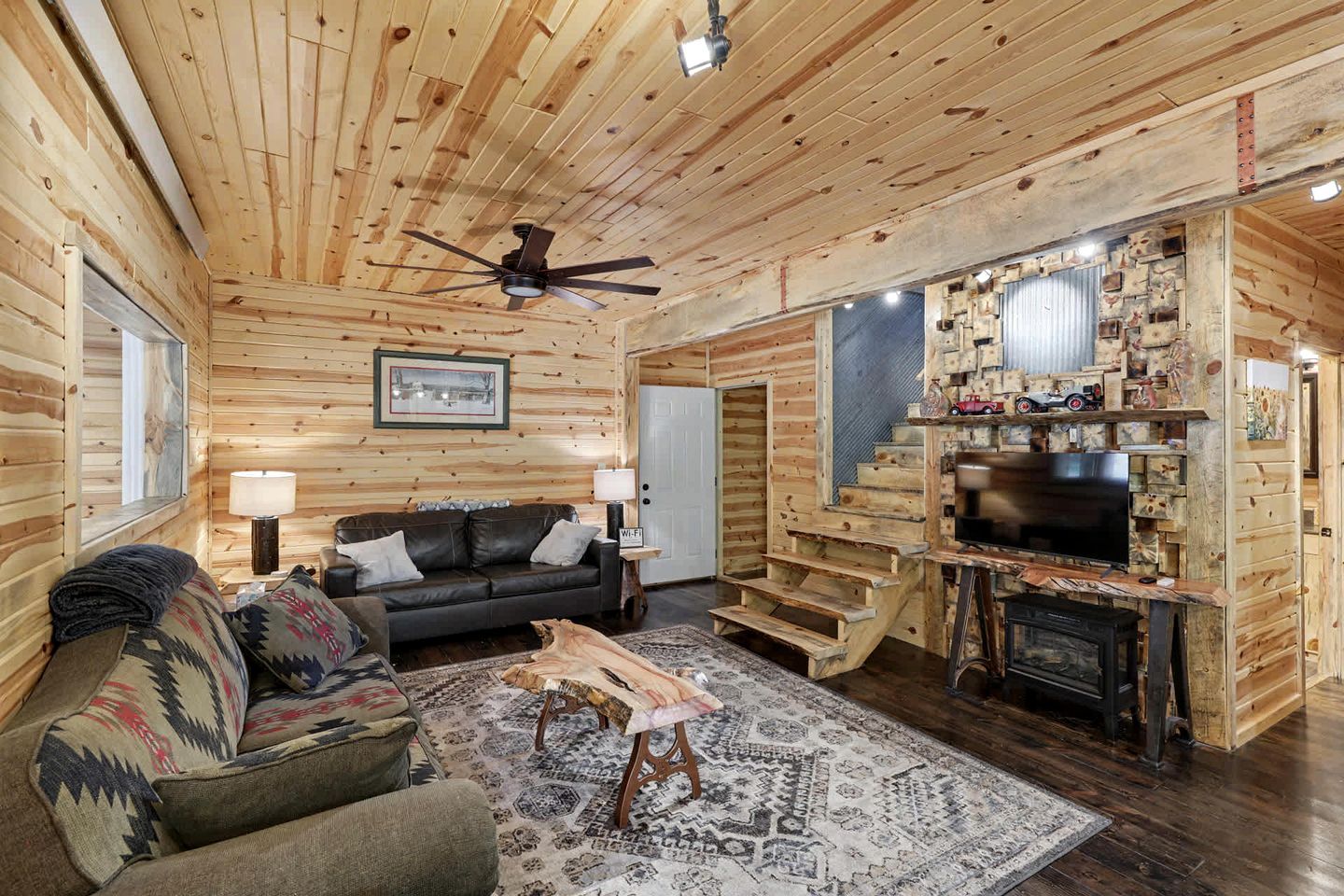 Interior de una cabaña de madera con sofá, mesa de centro, chimenea, televisor y escalera.