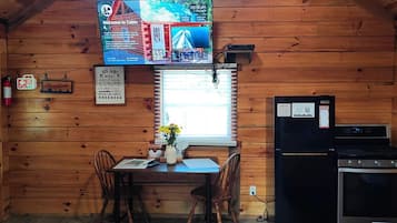 A cabin interior with a dining table, chairs, a small kitchen area with a stove, and a television mounted on the wall displaying a welcome message.