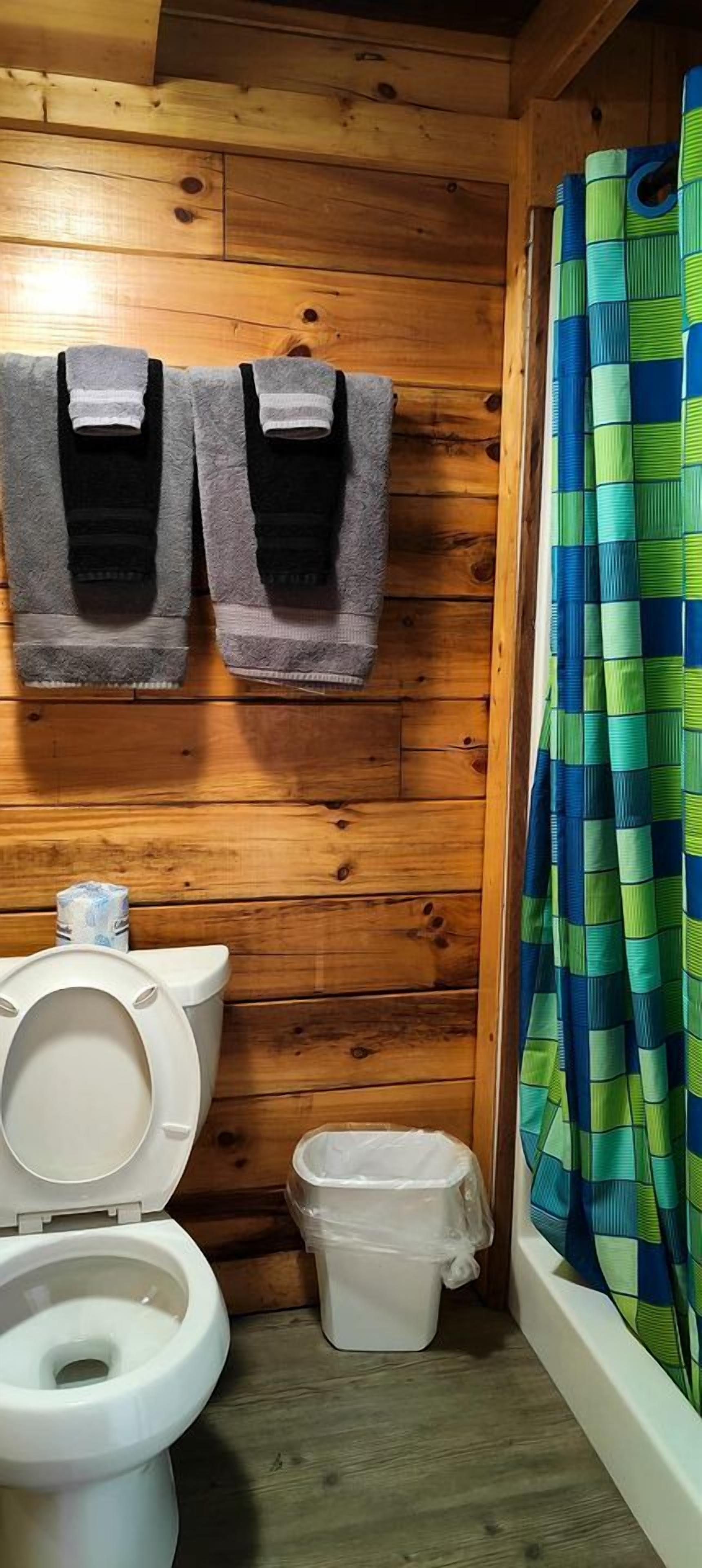 A wooden cabin bathroom with a toilet, two towels, a trash bin, and a shower curtain.