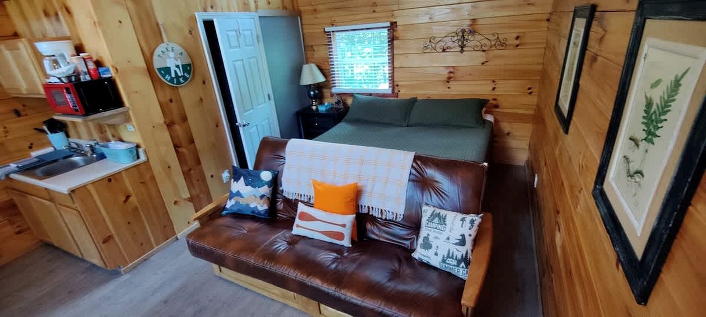 Ein gemütlicher Raum mit braunem Ledersofa, einer kleinen Küchenzeile, einem Bett mit grüner Bettdecke und einem Fenster mit Jalousien.