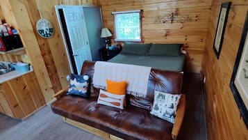 A compact room with a brown leather sofa, a bed with green bedding, a small kitchenette with a microwave, and a clock on the wall.