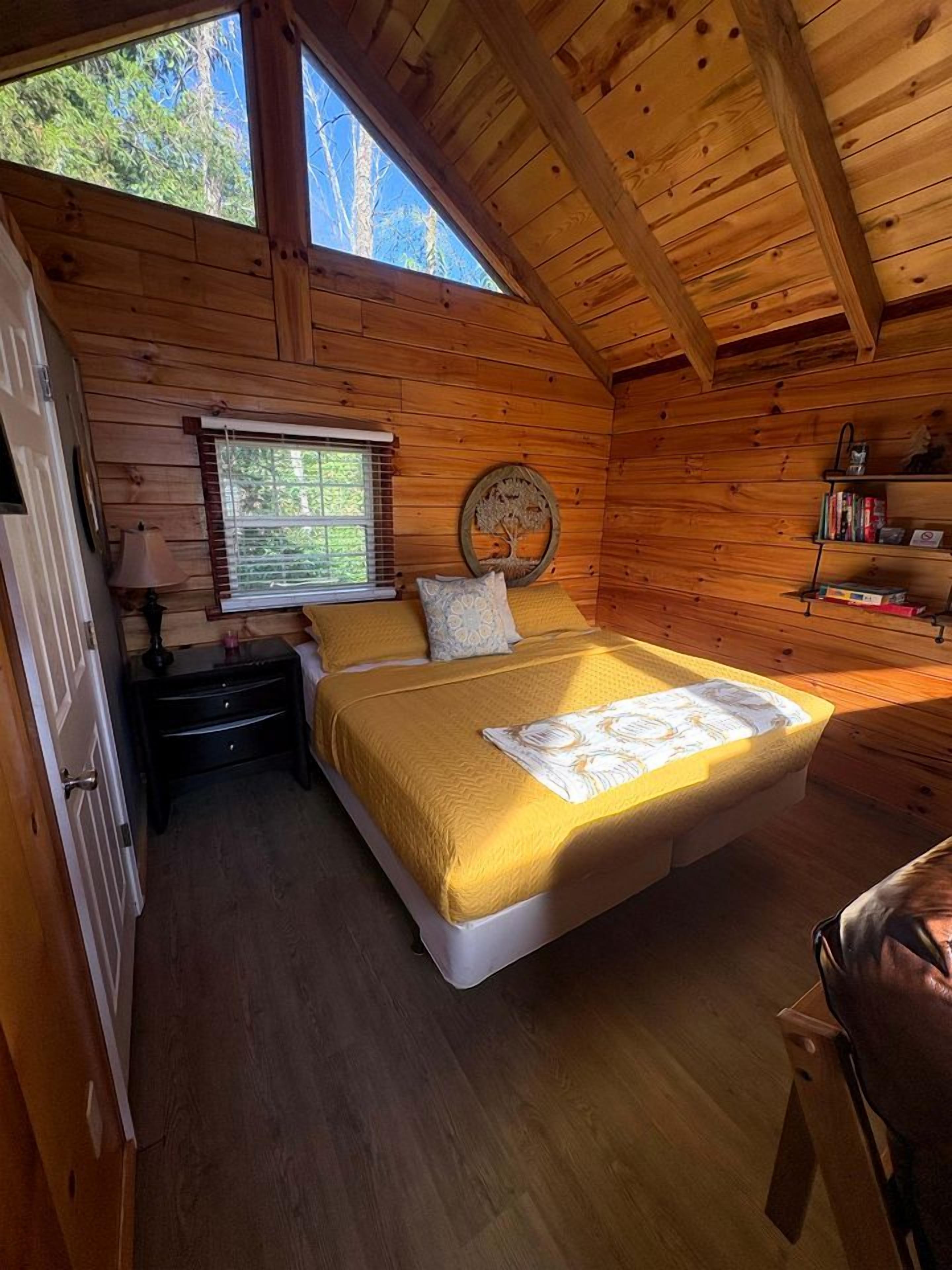 A wooden cabin bedroom with a bed, a nightstand, a window, and a shelf with books.