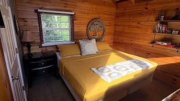 A wooden cabin bedroom with a bed, a nightstand, a window, and a shelf with books.