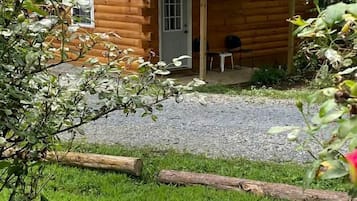A wooden cabin with a metal roof, a gravel driveway, and a grassy area with a pink flower in the foreground.