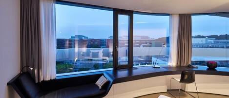 Una habitación moderna con un asiento curvo junto a la ventana, una silla negra y vistas a un paisaje urbano.