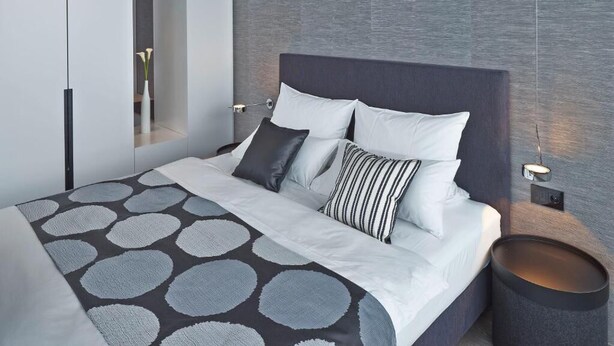 A modern bedroom with a large bed, a bedside table, a nightstand, and a wall-mounted lamp. - Campus at Home (Planegg)