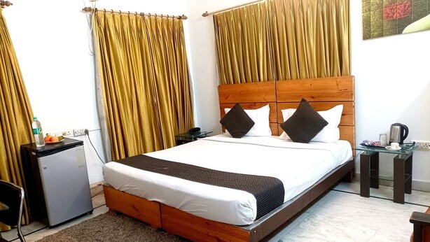 Una habitación de hotel con una cama de madera, un refrigerador pequeño, una mesita de vidrio y cortinas.