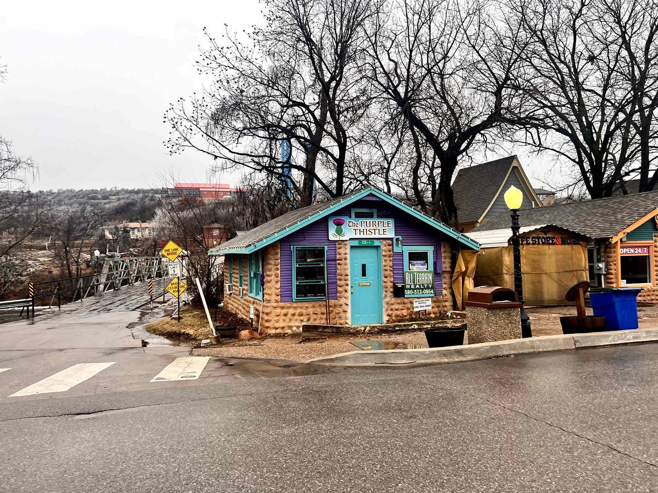 Un edificio pequeño con fachada púrpura y azul, un letrero que dice 'The Purple Thistle' y un escaparate con el letrero 'All Terrain Realty'.