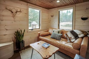 A cozy cabin interior with a leather sofa, a coffee table, and a large window offering a view of the forest.