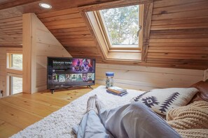 A cozy room with a sloped ceiling, a skylight, a television displaying a show, a bed with a patterned blanket, and a small table with books and a container.