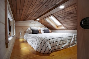 Une chambre avec un plafond en pente, des murs en bois, un puits de lumière, un lit avec une literie à rayures et une table de chevet.