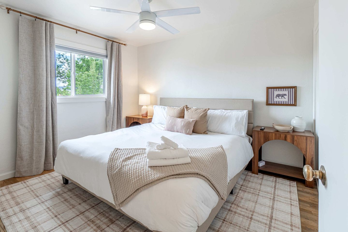 A neatly arranged hotel room with a bed, bedside tables, a lamp, a framed picture, and a view of greenery outside.