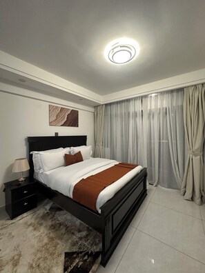 Un dormitorio moderno con cama, mesita de noche, lámpara, un cuadro en la pared y cortinas traslúcidas.
