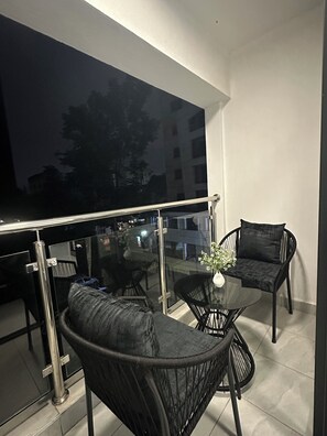 Un balcón con puerta de cristal, dos sillas de mimbre negro, una mesa pequeña con un florero y vistas a edificios y árboles.