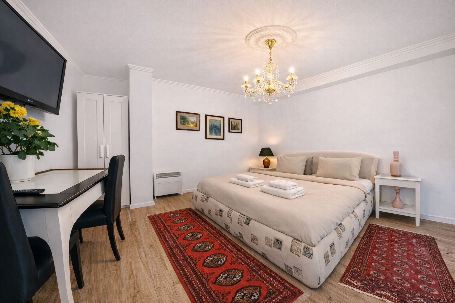 Ein Hotelzimmer mit Bett, Schreibtisch, Sessel, Fernseher, einer Kristallleuchtre und Bildern, sowie einem roten Teppich mit Muster.