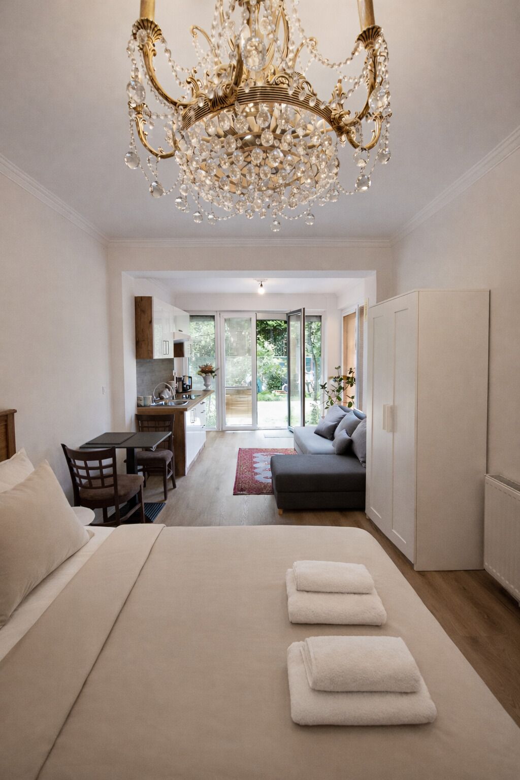 A modern bedroom with a large bed, a chandelier, a sofa, and a view of a kitchen and outdoor area.