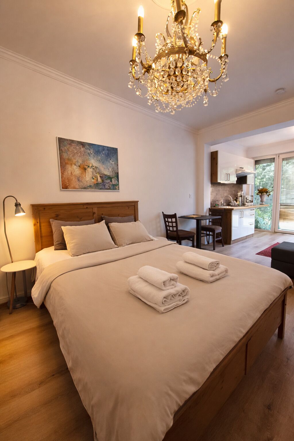 A hotel room with a large bed, a chandelier, a painting on the wall, and a view of the kitchen and outside.